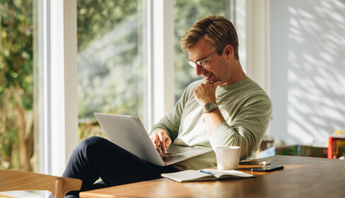 Man sits at dining room table researching homes for sale on his laptop.